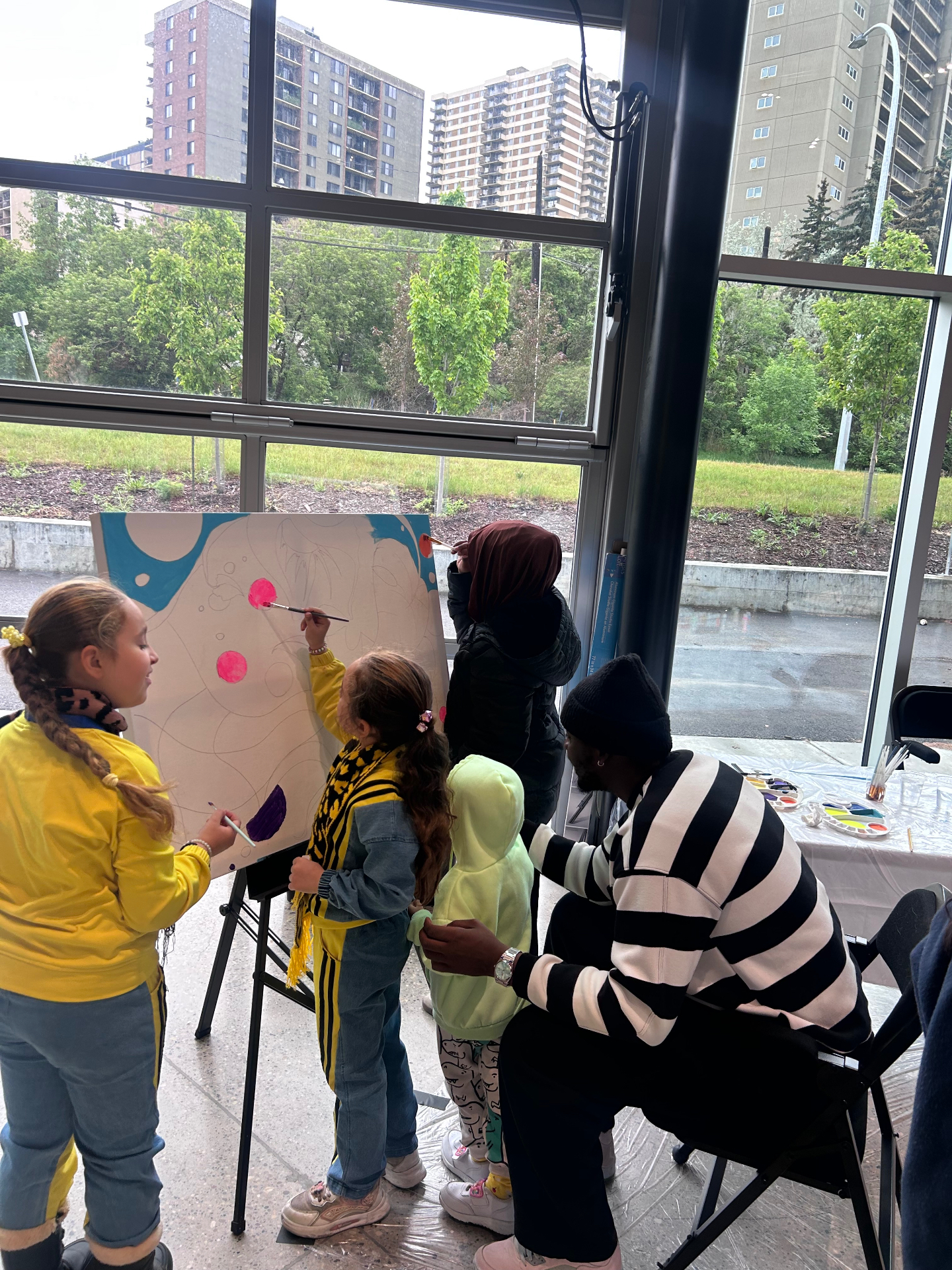 Children and a mentor painting a large collaborative canvas in front of bright studio windows