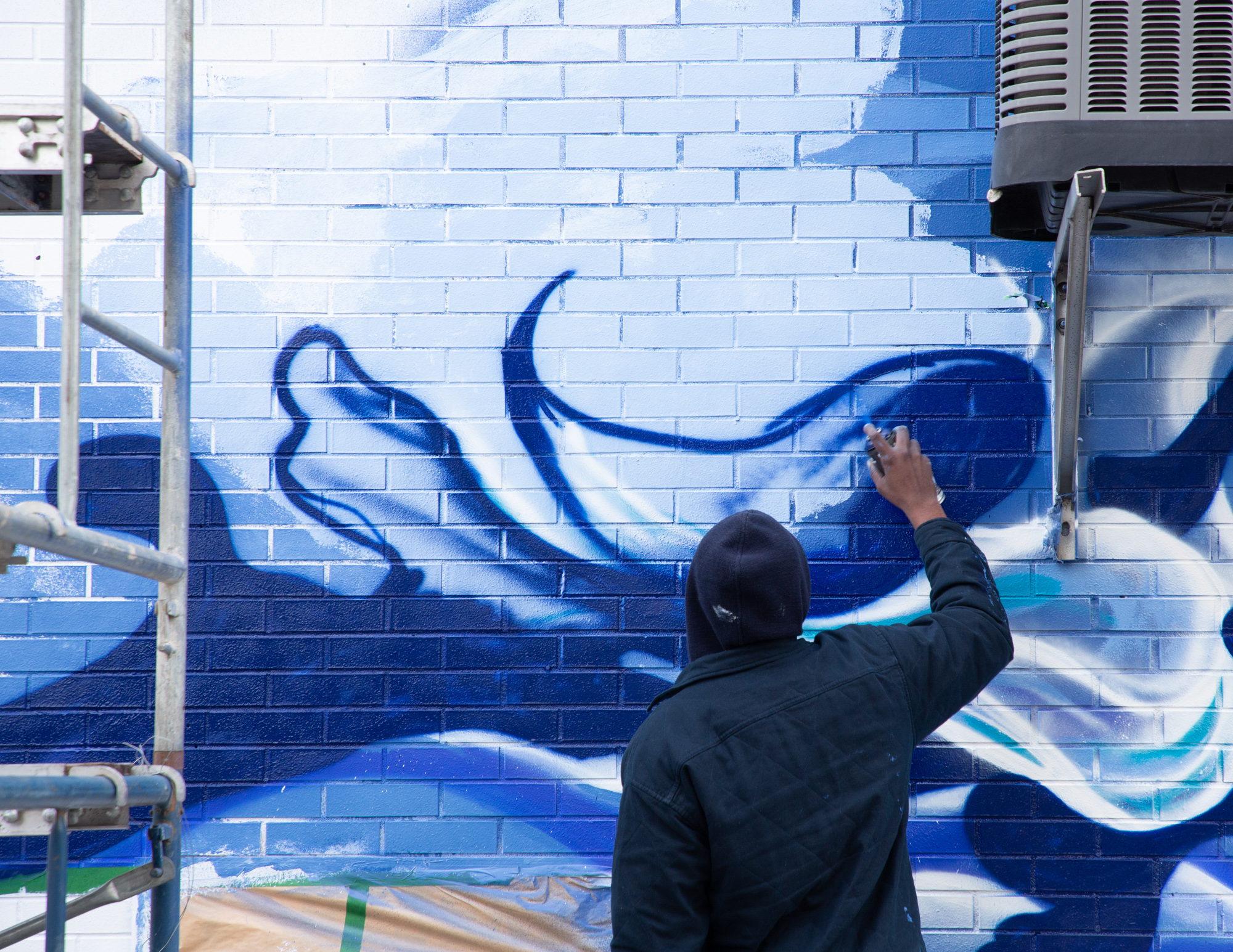 Dino, hooded, painting a large blue and white mural on a brick wall